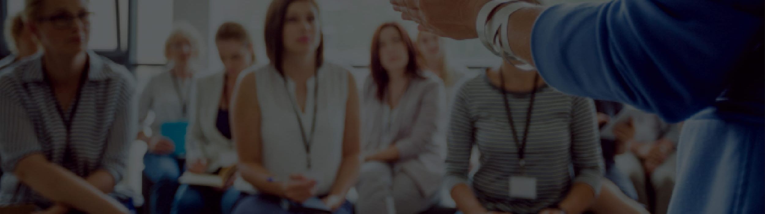 Close-up shot of a speaker's arm and hand gesturing during a presentation; in the background, an audience of seated people listens attentively in a professional training setting